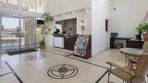 Een hotel lobby met een receptiebalie, een tijdschriftenhouder, een zithoek met rieten stoelen en een grote glazen deur die naar buiten leidt.