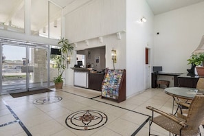 Vestíbulo de hotel con mostrador de recepción, un atril con revistas, una zona de estar con sillones de mimbre y un gran ventanal que da al exterior.