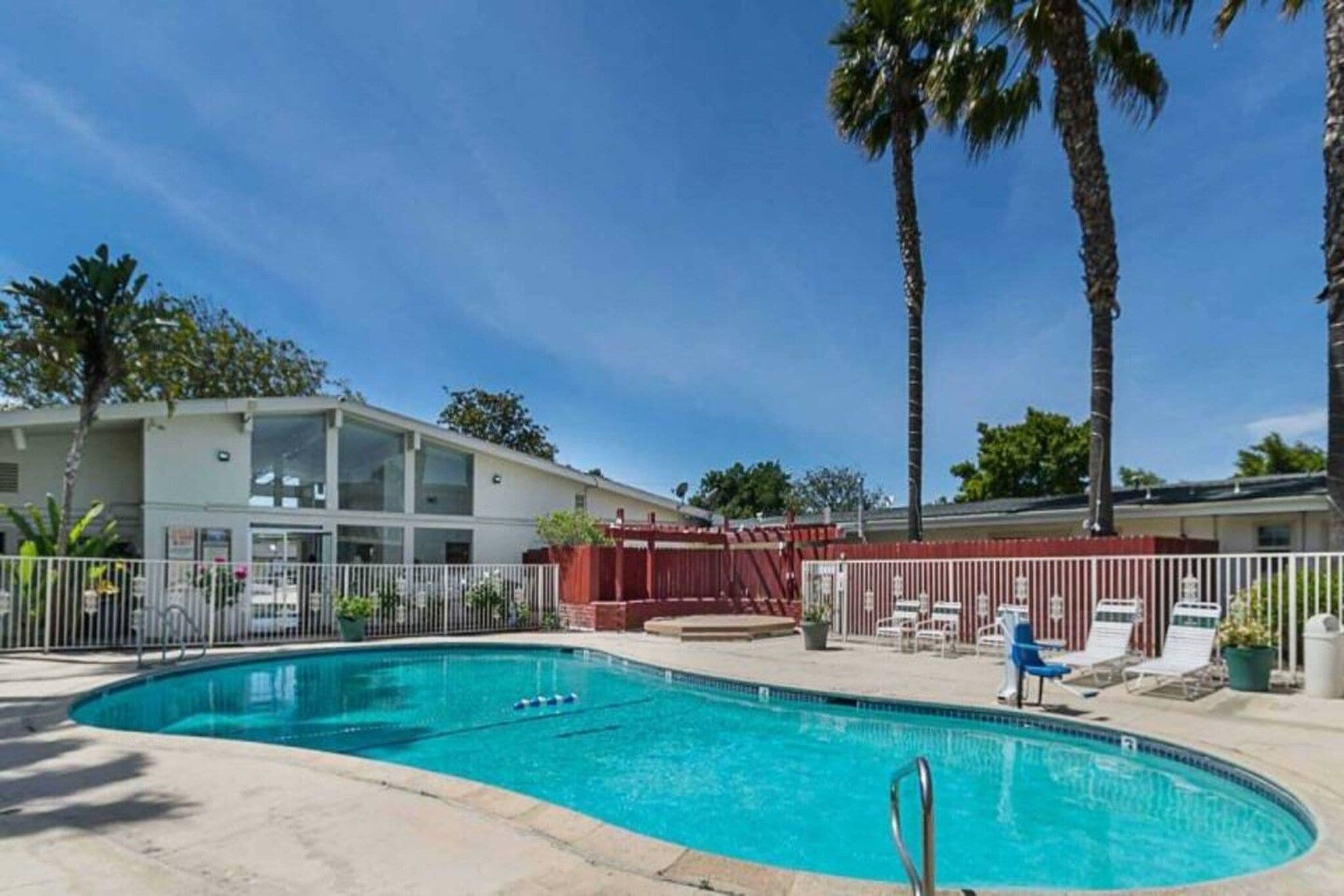 A swimming pool with a fenced area, palm trees, and a building with a white facade.