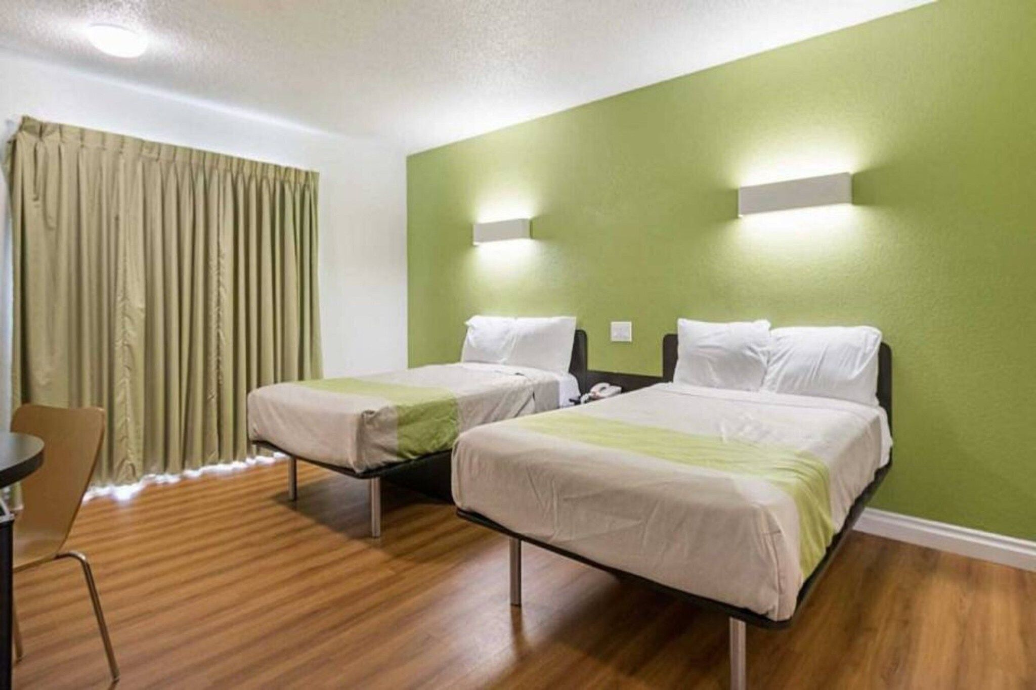 A hotel room with two beds, a wooden floor, a green wall, and a window with curtains.