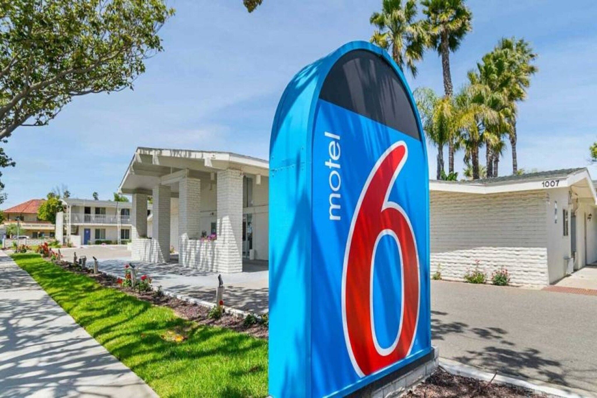 Placa de motel com o número 6, estacionamento e um prédio de fachada branca.