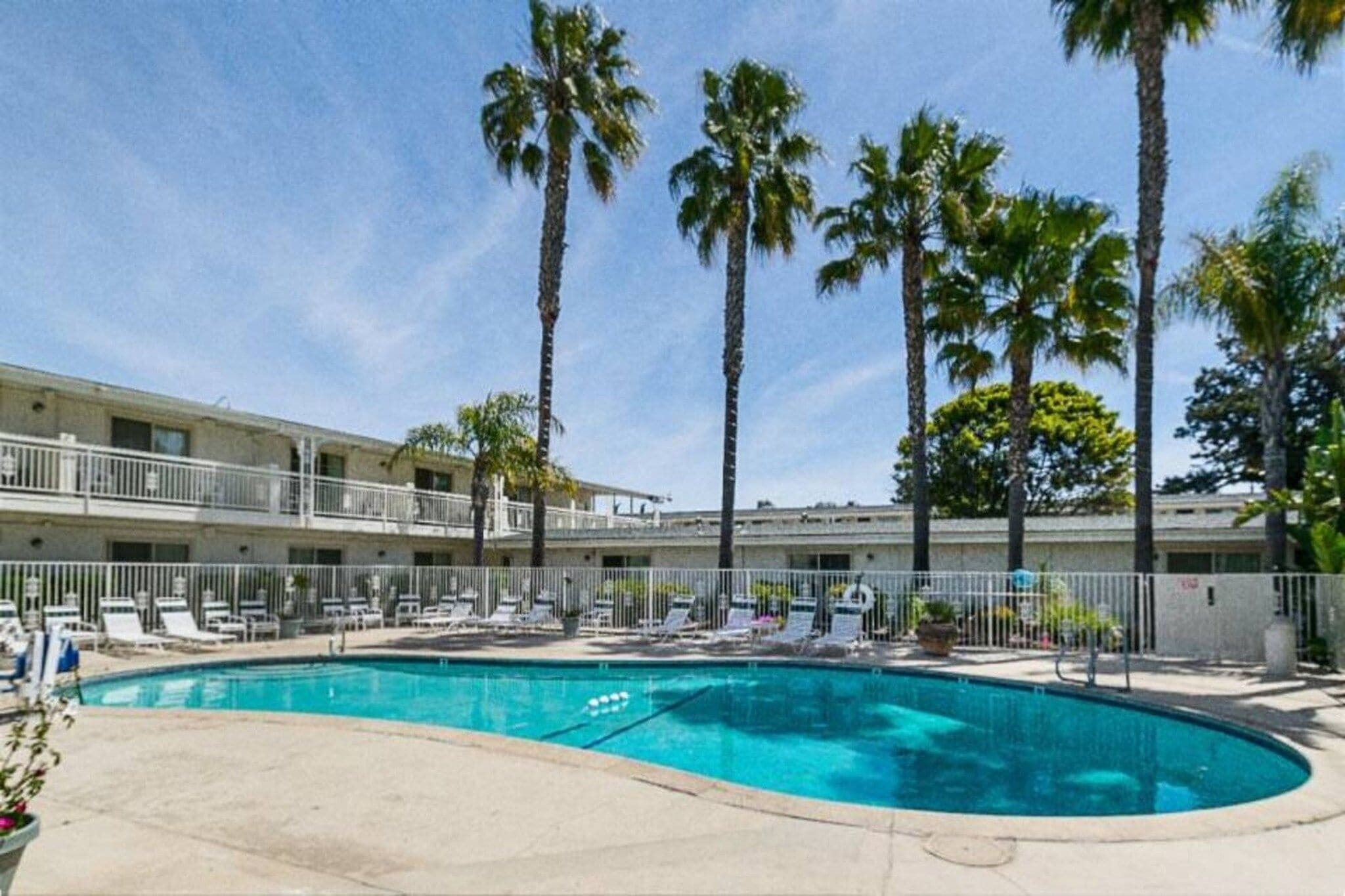 A swimming pool with lounge chairs, palm trees, and a multi-story building with balconies.