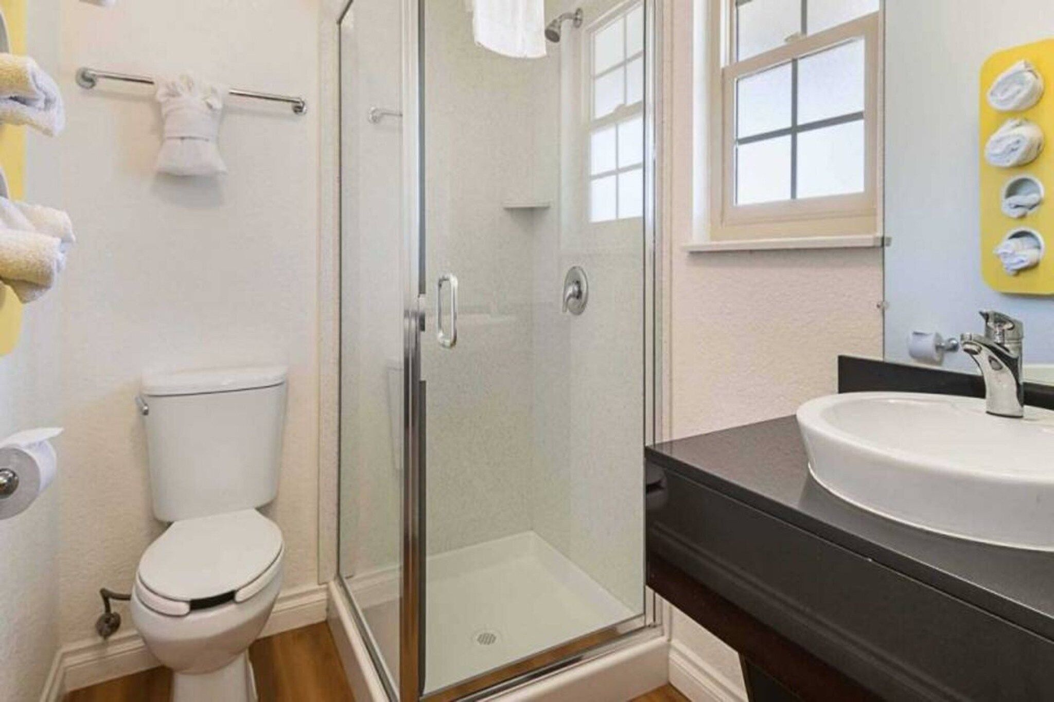 Et badeværelse med en glasafskærmet brusekabine, et hvidt toilet og en vask med en mørk bordplade.