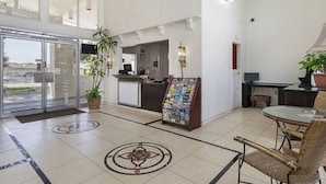 Een hotel lobby met een receptiebalie, een tijdschriftenhouder, een zithoek met rieten stoelen en een grote glazen deur die naar buiten leidt.