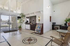 Vestíbulo de hotel con mostrador de recepción, un atril con revistas, una zona de estar con sillones de mimbre y un gran ventanal que da al exterior.