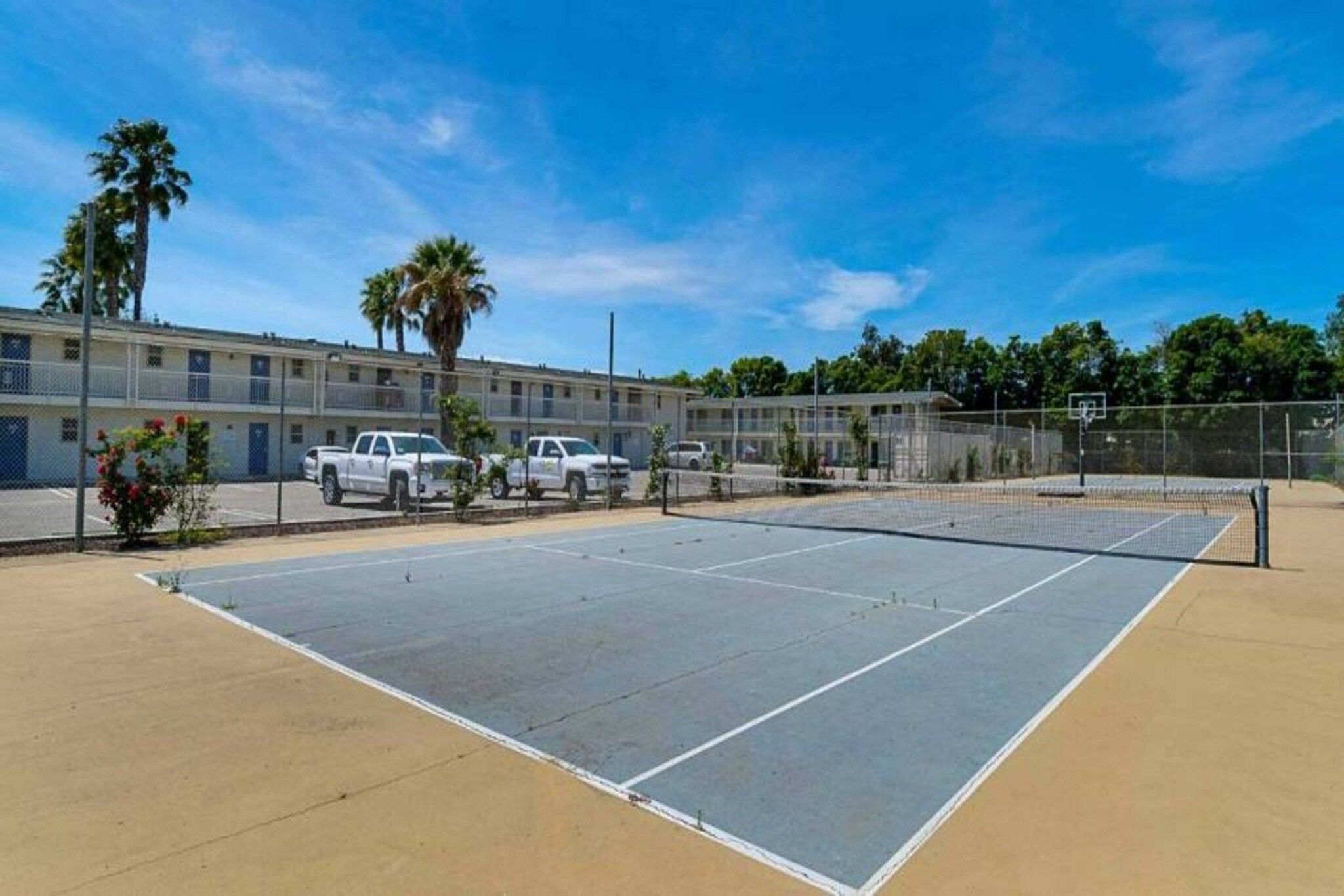 Una cancha de tenis al aire libre con red, rodeada por una cerca y contigua a un edificio de varios pisos con vehículos estacionados.