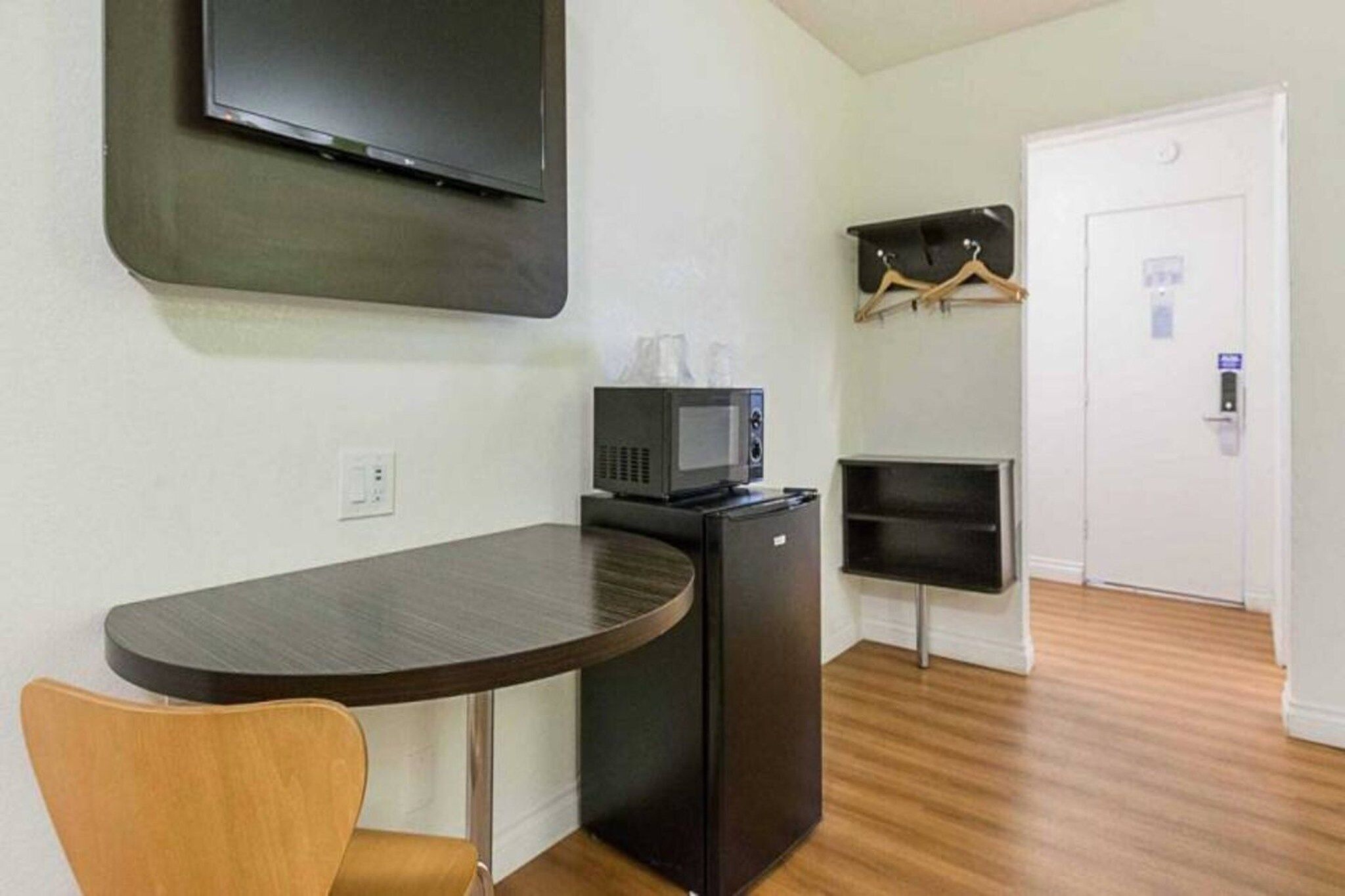 Habitación compacta con televisión de pantalla plana en la pared, un refrigerador pequeño, un microondas y una silla de madera.