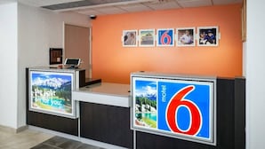 Una recepción con un panel digital que muestra un mensaje panorámico, el número '6' que identifica al motel, y una fila de fotografías enmarcadas en la pared.
