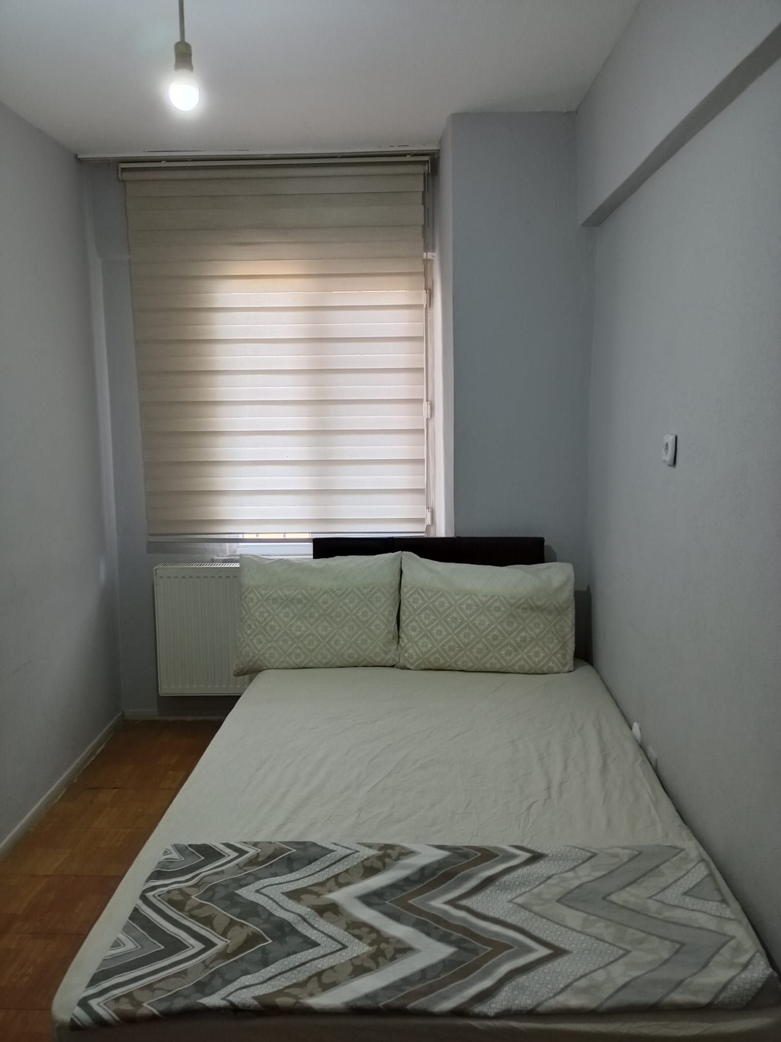 Un dormitorio pequeño con una cama individual, un radiador y una ventana con persianas.