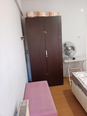 Ein Zimmer mit einem Bett, einem Schreibtisch mit Ventilator, einem Kleiderschrank und einer Heizung.