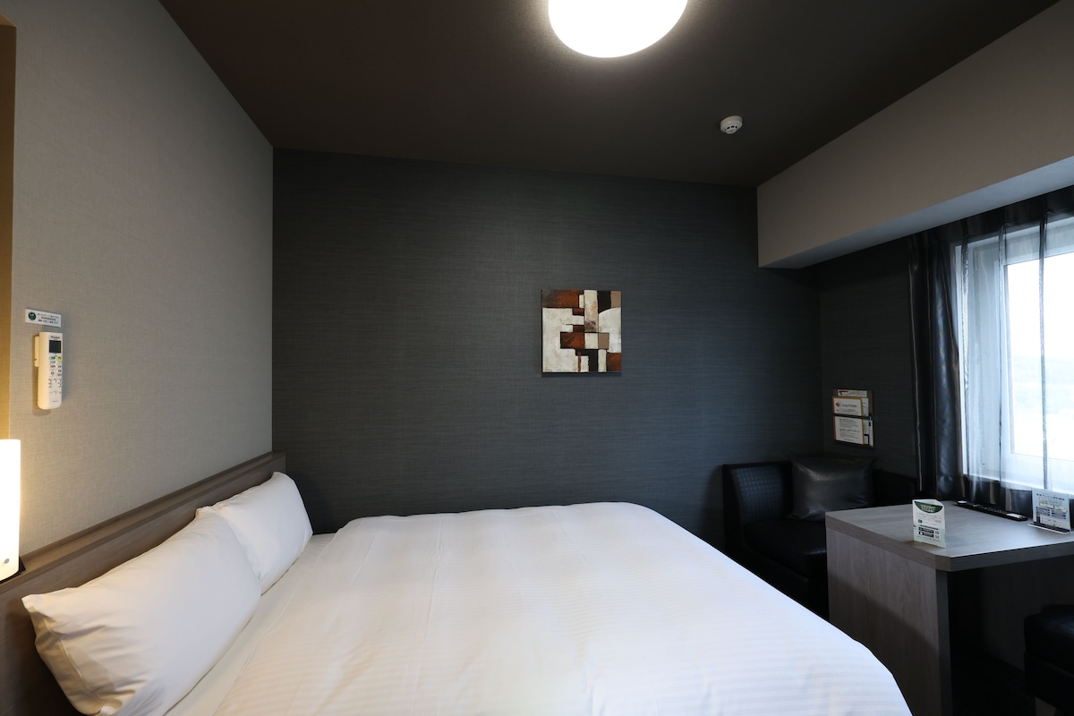 A hotel room with a large bed, a desk, and a chair. There is a painting on the wall and a window with curtains.