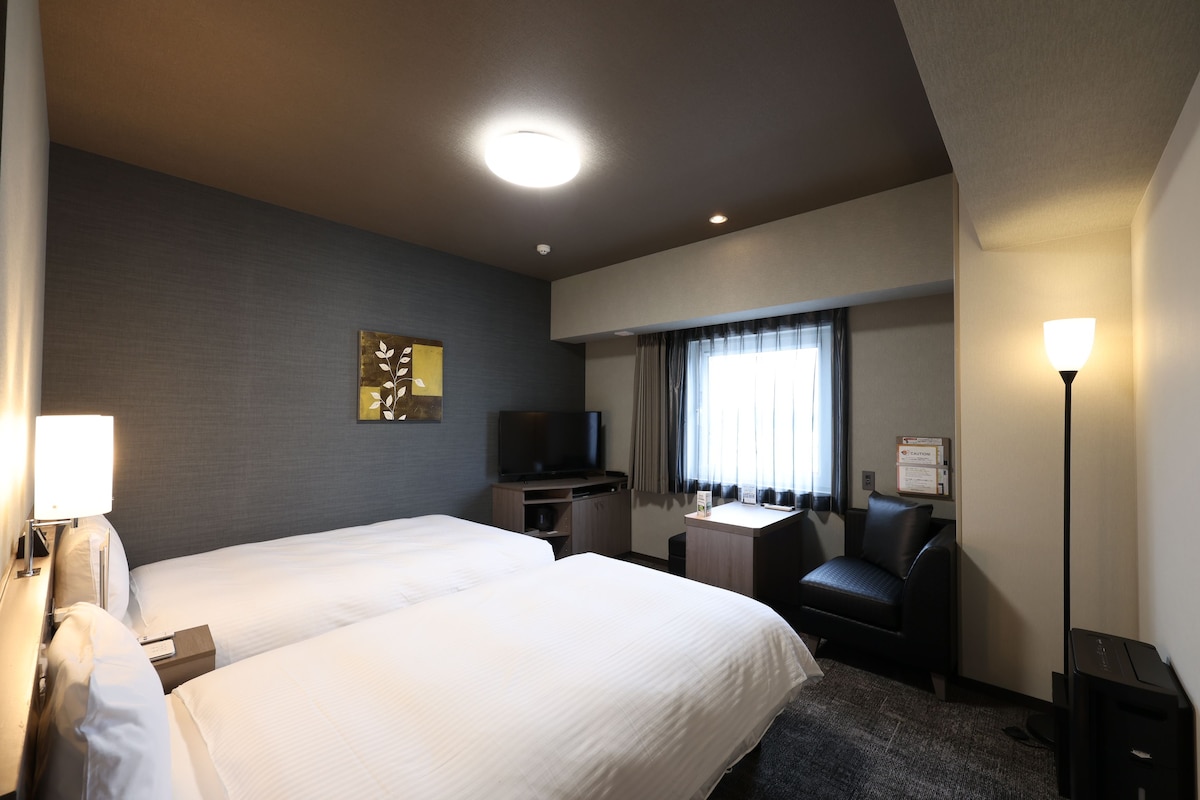 A hotel room with two beds, a desk, a television, a chair, and a lamp.