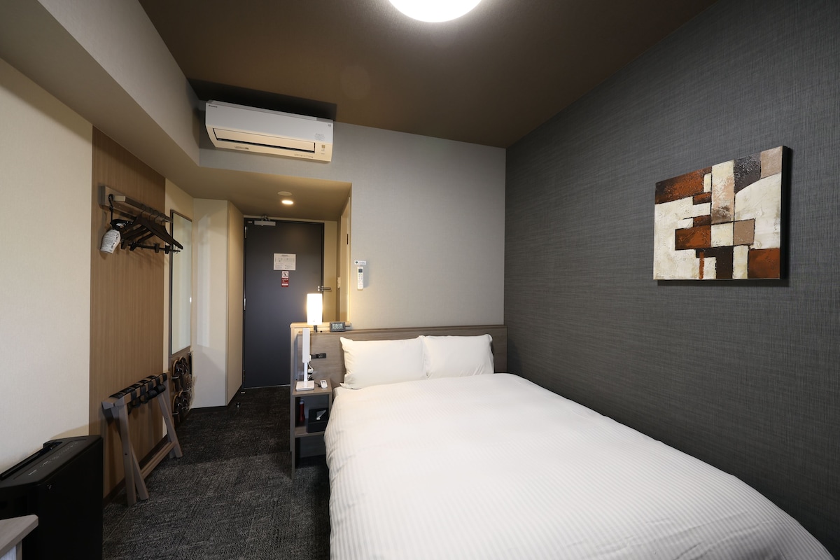 A hotel room with a bed, a desk, a chair, an air conditioning unit, a painting on the wall, and a door leading to another room.