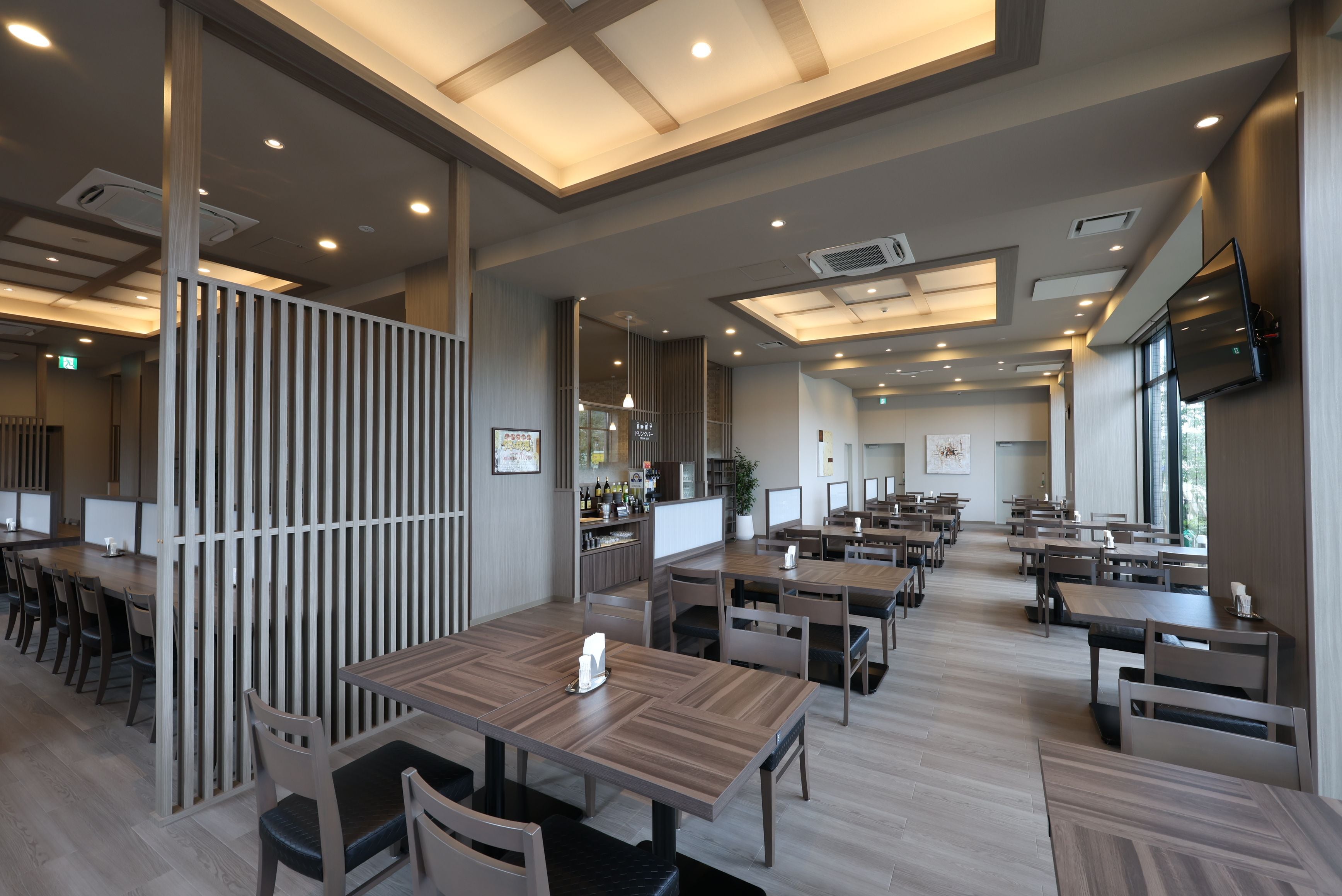 A modern restaurant interior with multiple dining tables and chairs, recessed ceiling lighting, and a flat-screen TV mounted on the wall.