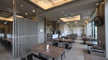 A modern restaurant interior with multiple dining tables and chairs, recessed ceiling lighting, and a flat-screen TV mounted on the wall.