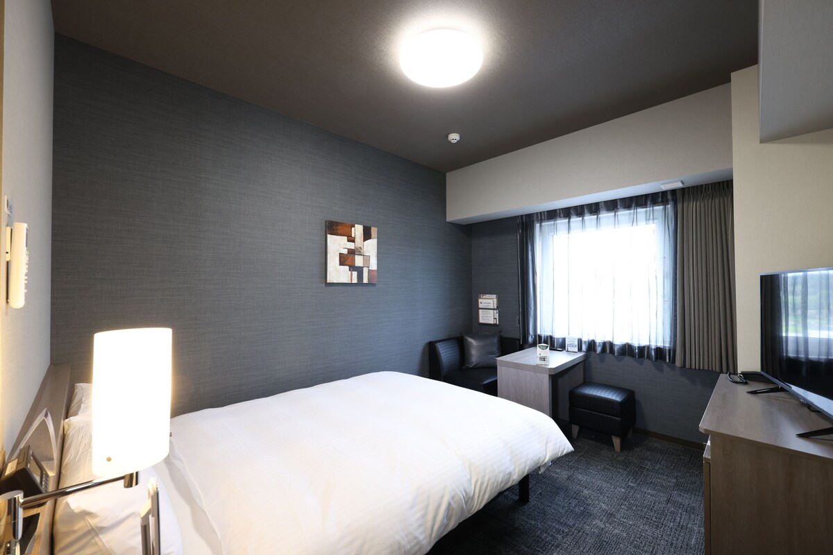 A hotel room with a bed, a desk, a chair, a television, and a window with curtains.