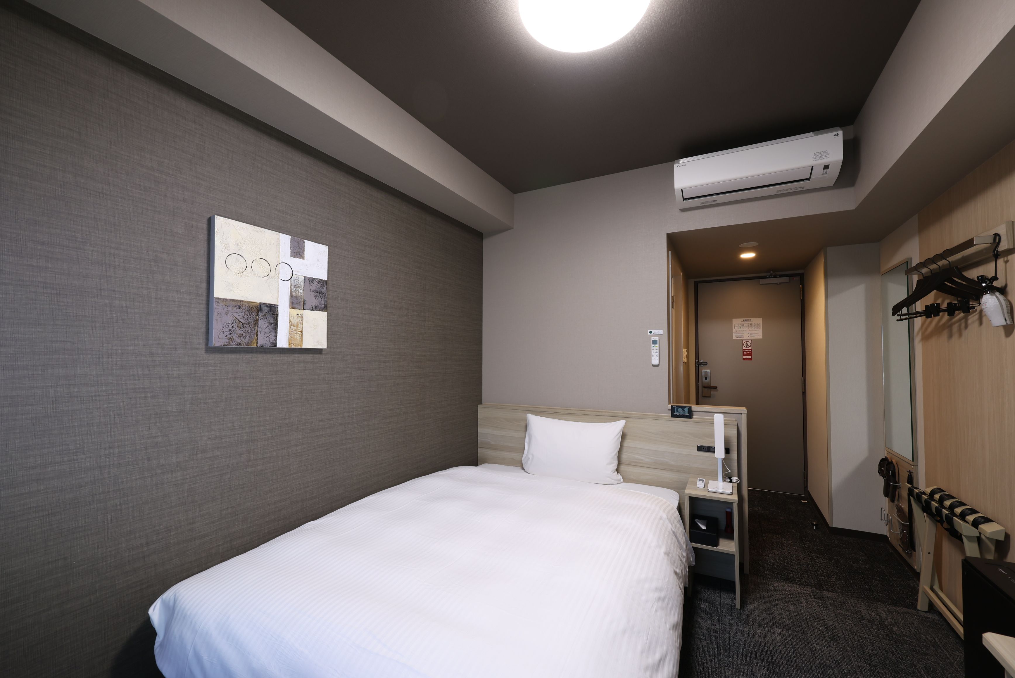A hotel room with a bed, a wall-mounted air conditioner, a small bedside table, a coat rack, and a door leading to a hallway.