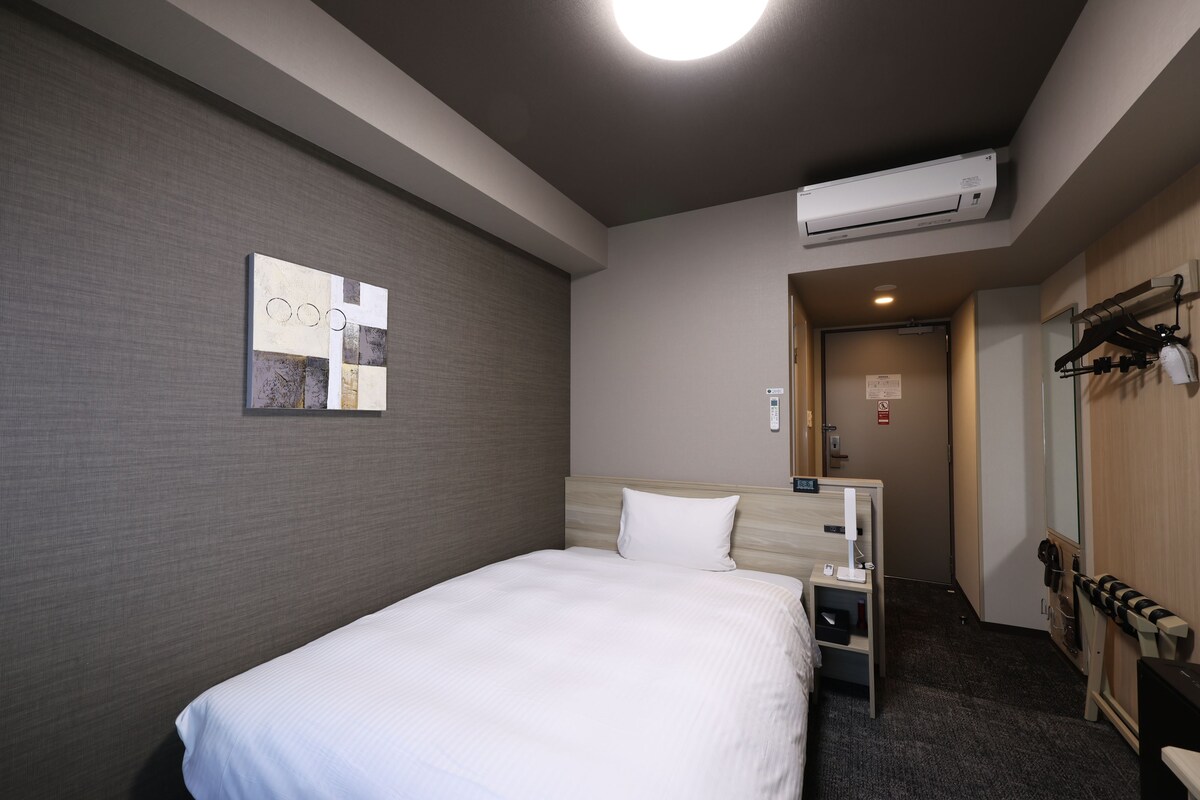 A hotel room with a bed, a wall-mounted air conditioner, a small bedside table, a coat rack, and a door leading to a hallway.