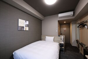 Habitación de hotel con cama, aire acondicionado de pared, mesita de noche pequeña, perchero y puerta que da a un pasillo.