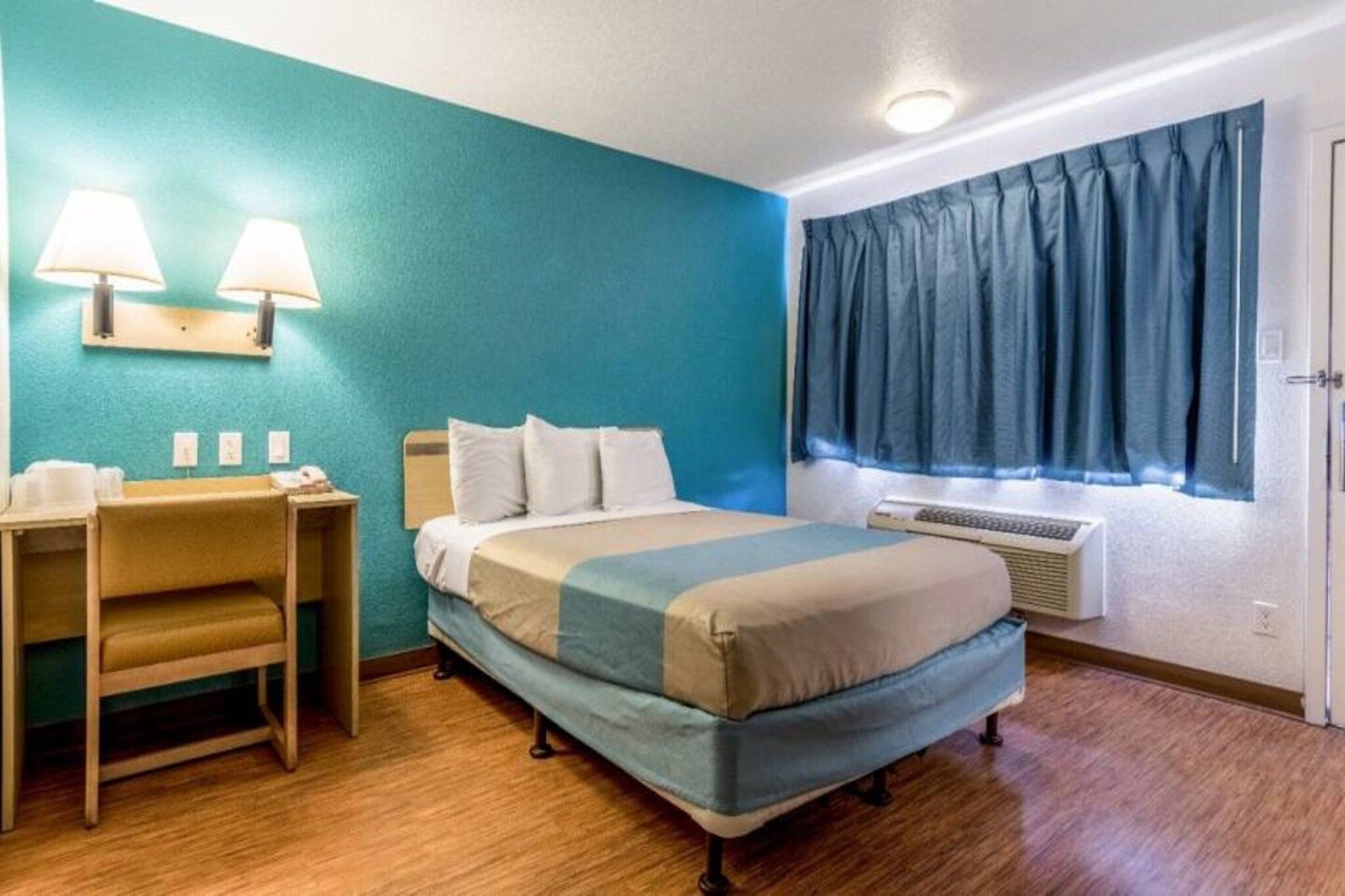 A hotel room with a bed, a desk, two lamps, a window with curtains, and an air conditioning unit.