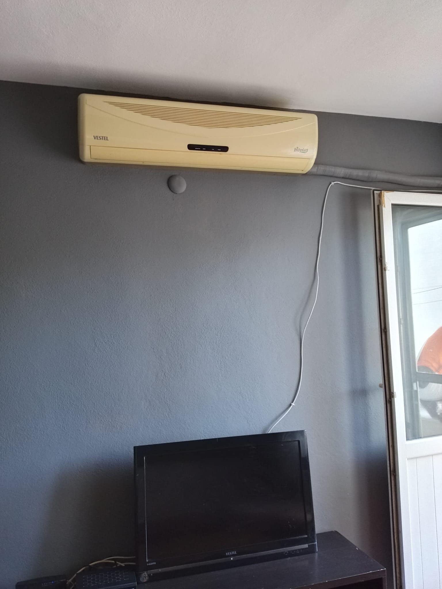 A room with a VESTEL air conditioner mounted on the wall, a black TV on a stand, and a window with a view.