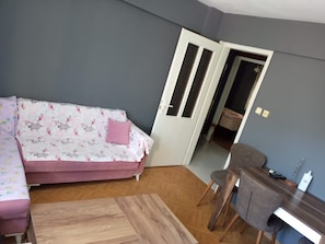 Ein Zimmer mit einer Couch, einem Holztisch, einem Schreibtisch mit Stuhl und einer Tür zu einem weiteren Raum.