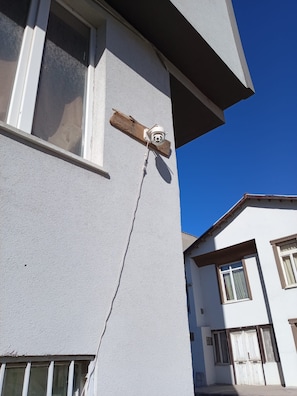A building corner with a white exterior, a window, and a security camera mounted on a wooden beam.