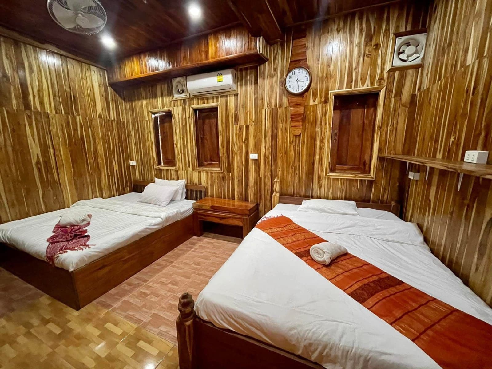 A room with two wooden beds, a wooden floor, a wall clock, and a ceiling fan.