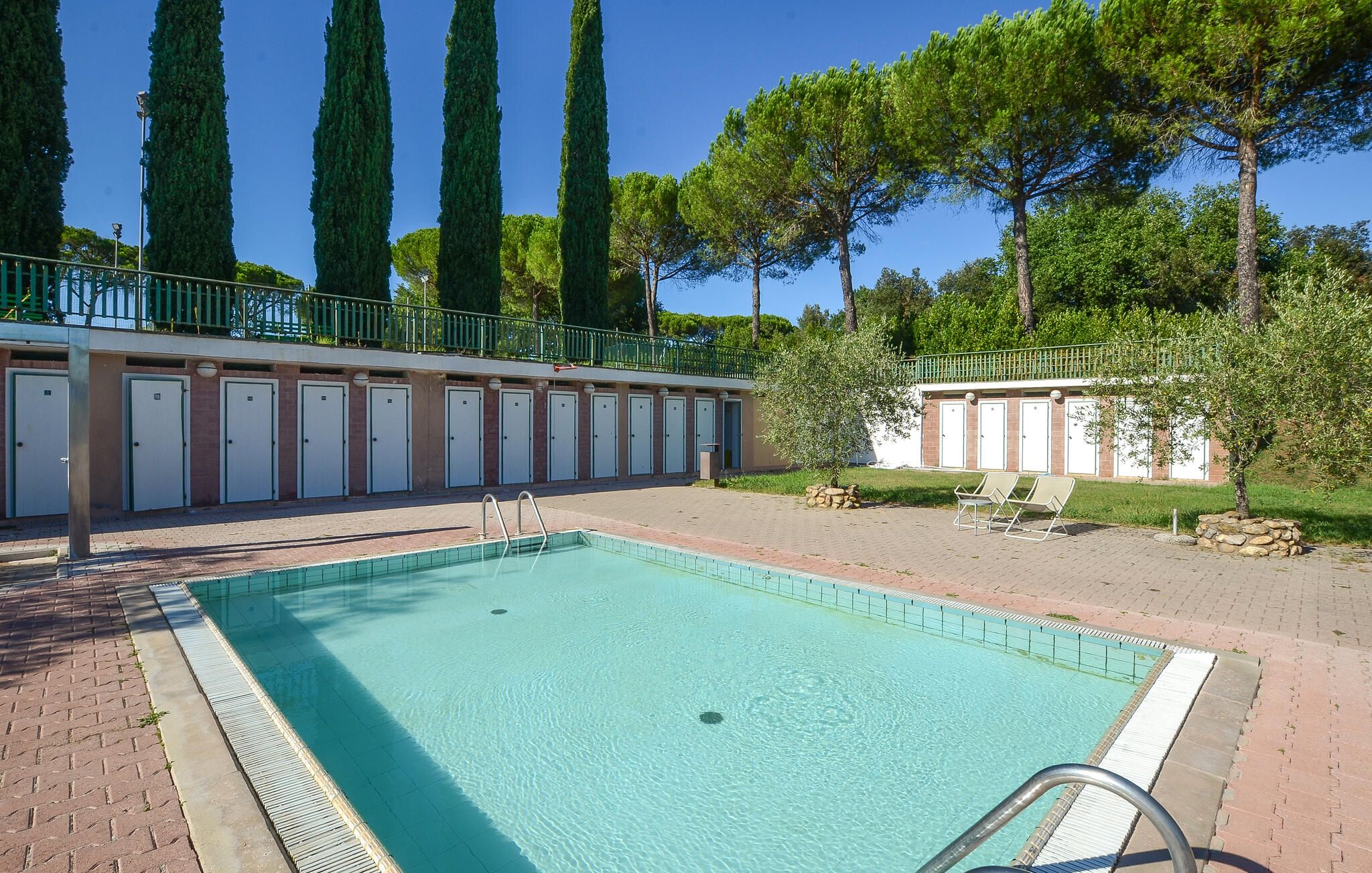 A swimming pool with a tiled deck, a row of doors, and a green fence.
