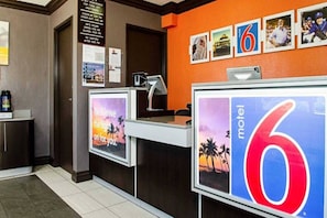 Mostrador de recepción de hotel con un cartel grande con el número '6', una computadora y un cartel con un atardecer y palmeras.