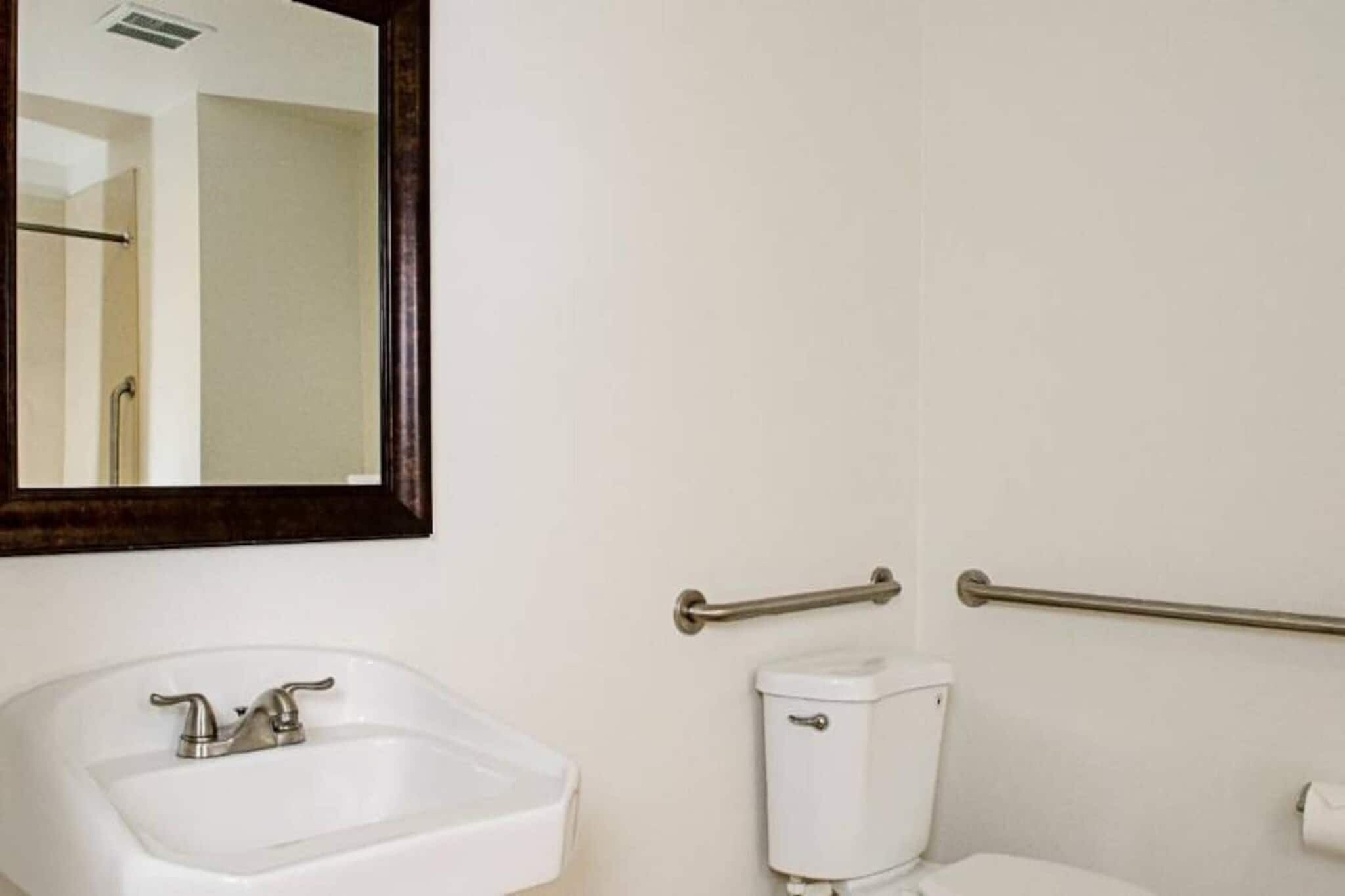 A bathroom with a white sink, a mirror, a toilet, and grab bars.
