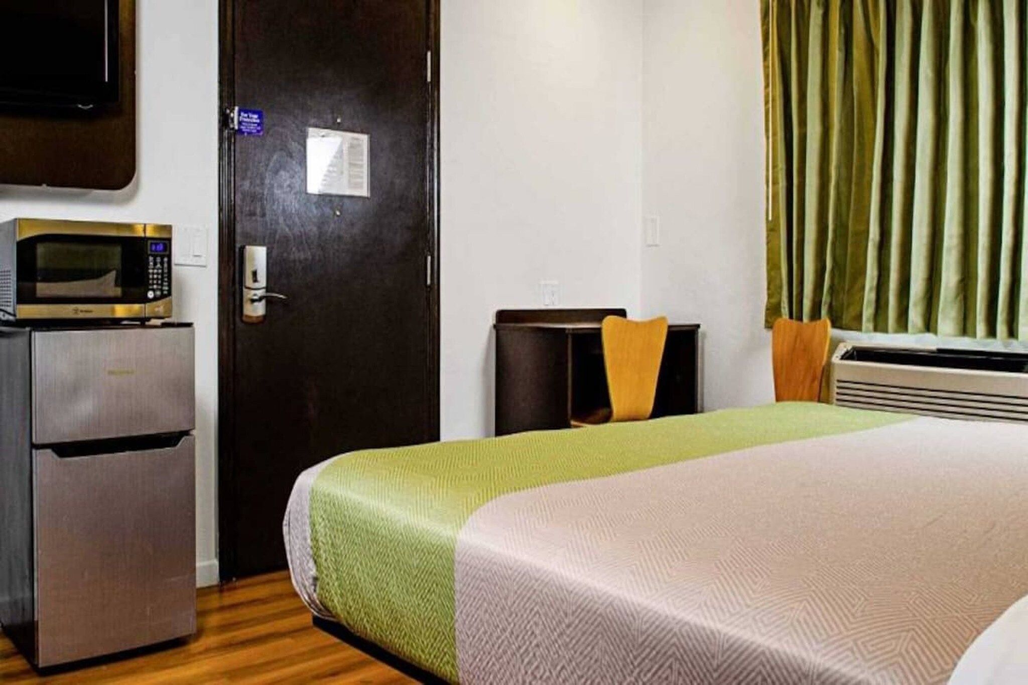 Une chambre d’hôtel avec un lit, un four à micro-ondes, un réfrigérateur, une porte avec une pancarte et une fenêtre avec des rideaux.