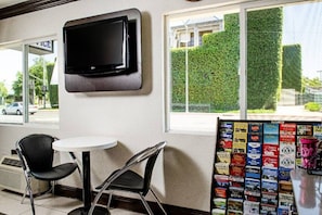 Habitación con televisor de pantalla plana montado en la pared, dos sillas negras alrededor de una pequeña mesa blanca y una ventana con vistas a un seto exterior.