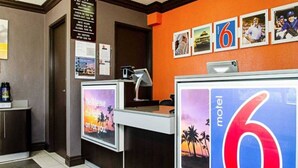 Mostrador de recepción de hotel con un cartel grande con el número '6', una computadora y un cartel con un atardecer y palmeras.