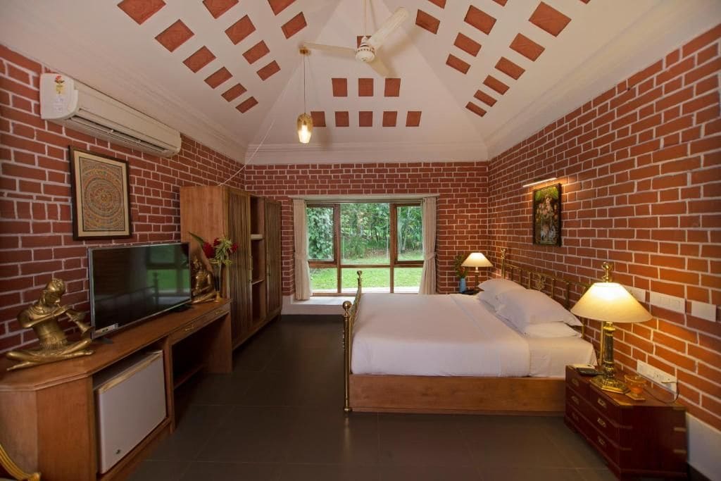 Quarto de hotel com parede de tijolos, cama, TV, escrivaninha e janela com vista para o verde.