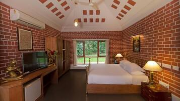 Quarto de hotel com parede de tijolos, cama, TV, escrivaninha e janela com vista para o verde.