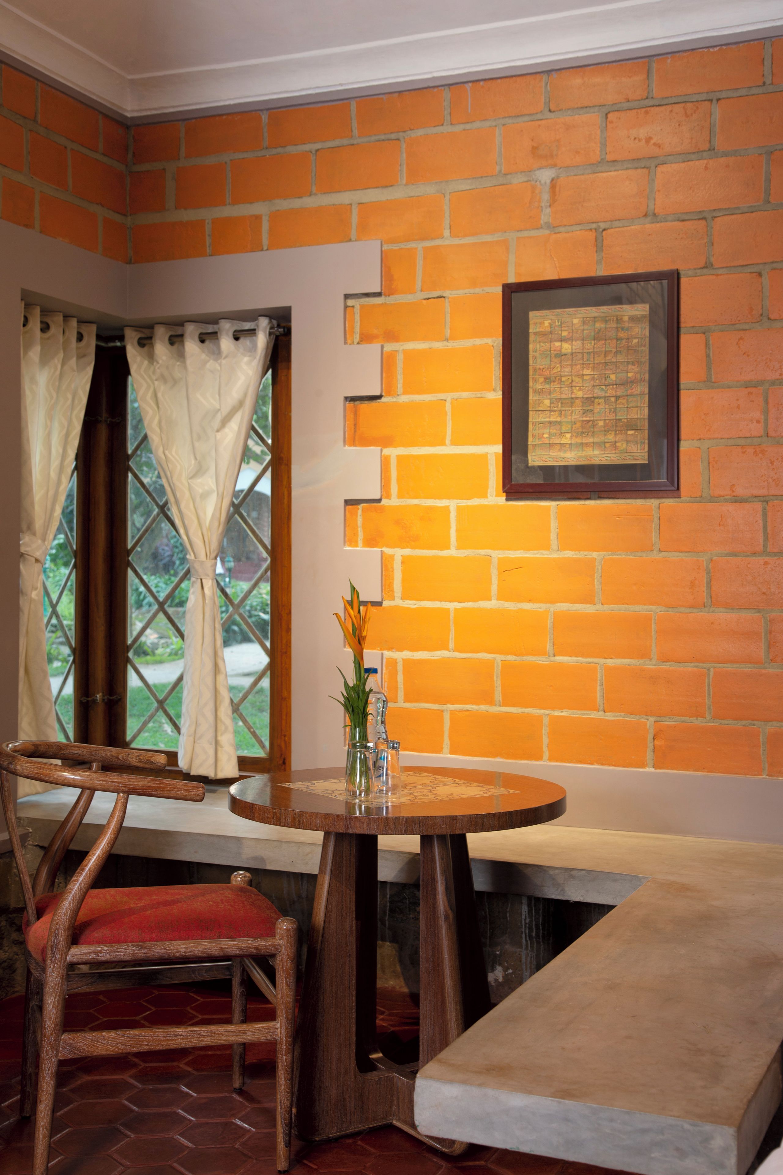 Una habitación de esquina con pared de ladrillo, una ventana con cortinas, una silla de madera, una mesa redonda de madera con un jarrón y un banco.