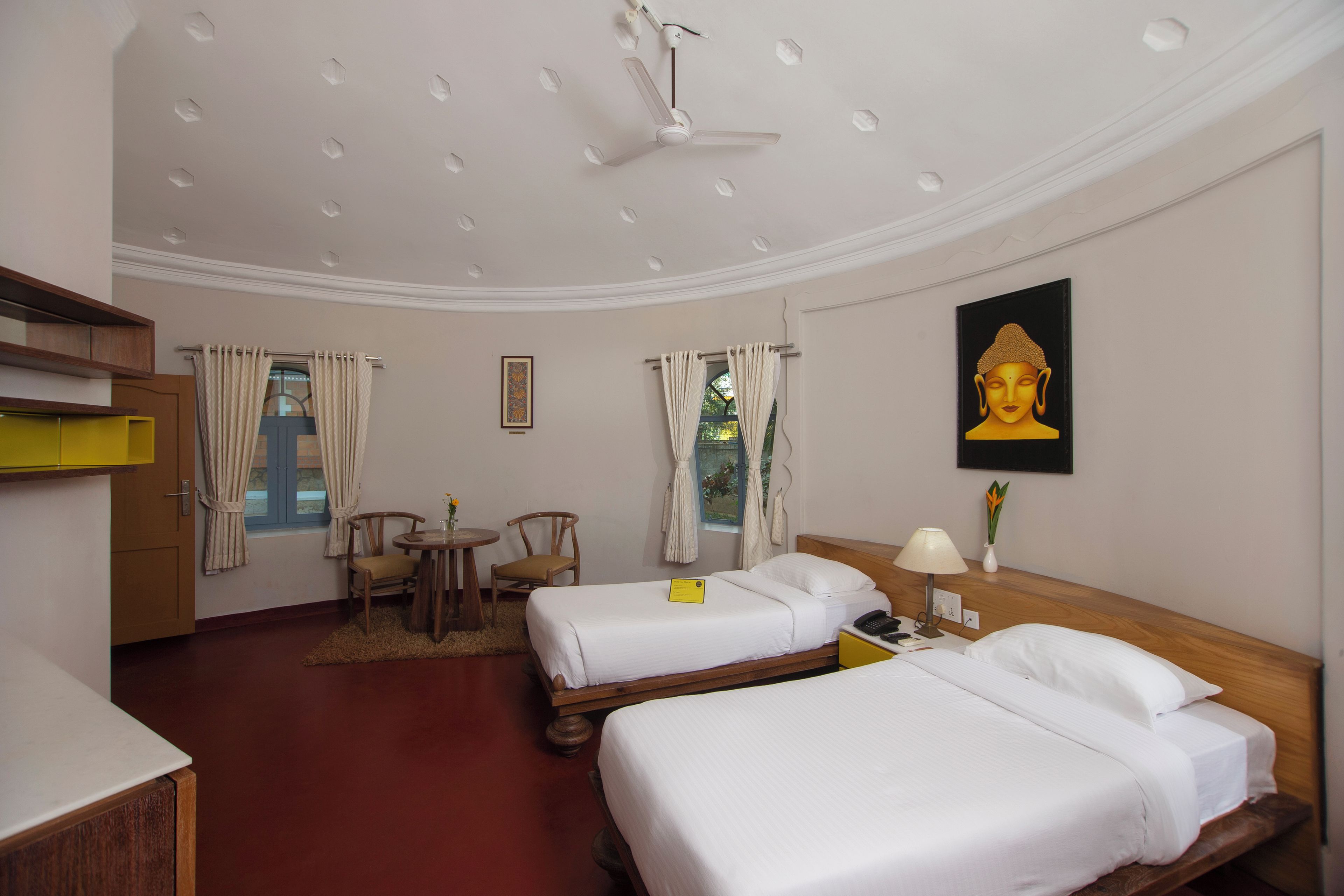 Habitación de hotel con dos camas, un escritorio, una silla, un ventilador de techo y una estatua de Buda.