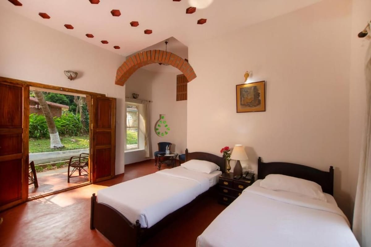 A room with two beds, a wooden door leading to a balcony, a small table with a chair, and a wall-mounted picture.
