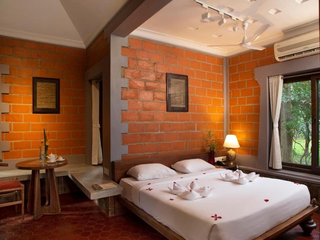 Habitación de hotel con pared de ladrillo, cama con ropa blanca, mesita de noche con lámpara y ventana con cortinas.