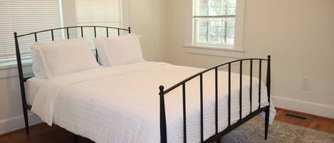 A single bed with a black metal frame, white bedding, and a patterned rug in a room with white walls and windows.
