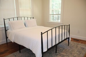 Un lit simple avec un cadre en métal noir, une literie blanche et un tapis à motifs, dans une pièce aux murs et aux fenêtres blancs.