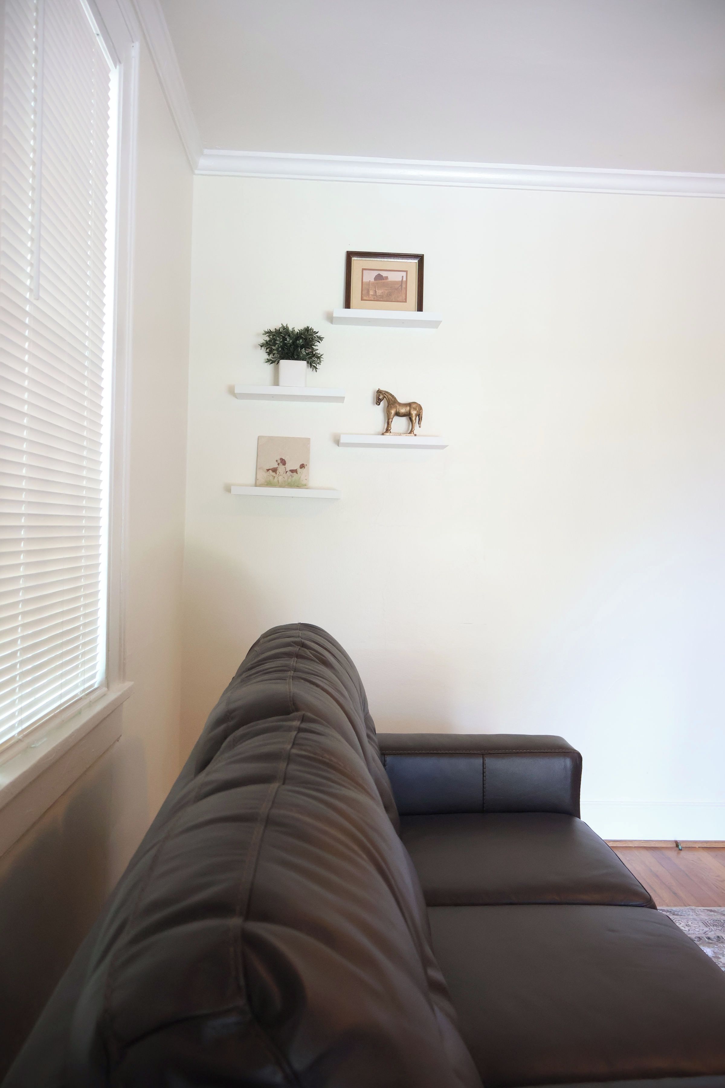 Een woonkamer met een bruine bank, witte muren en drie rekken met decoratieve voorwerpen.