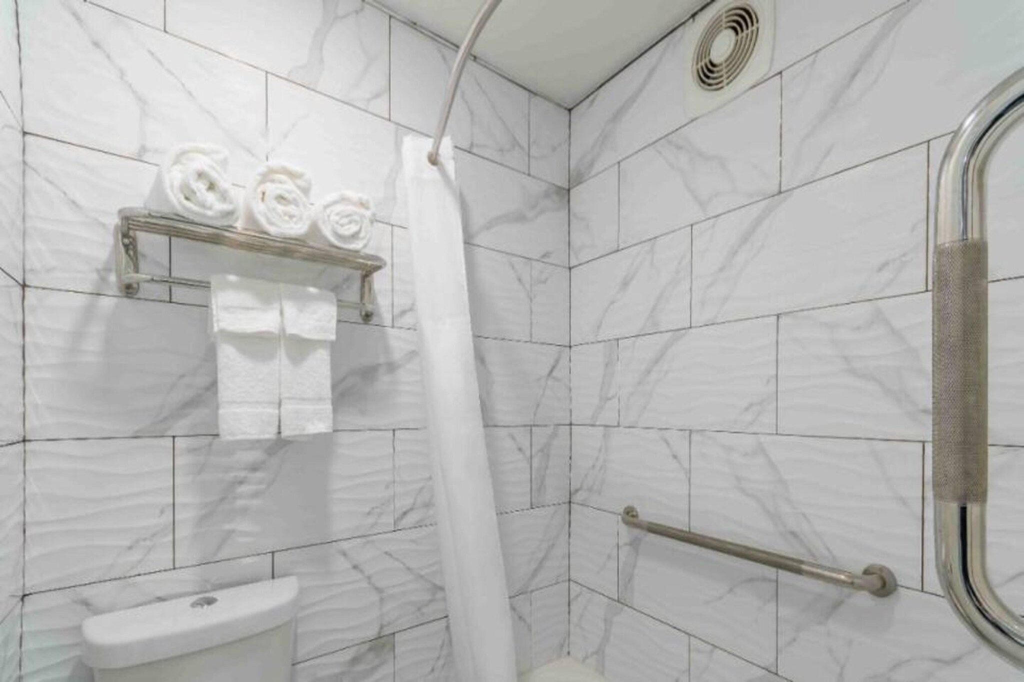 A bathroom with white marble tiles, a shower with a white curtain, a towel rack with rolled towels, and a handrail.
