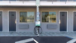 Un edificio con dos puertas identificadas con los números 106 y 105, una estación de carga para vehículos eléctricos y un aparcamiento con plazas delimitadas.