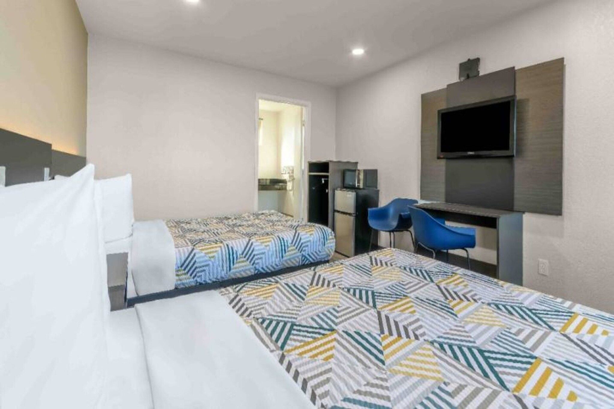 A hotel room with two beds, a desk, a chair, a TV, and a kitchenette.