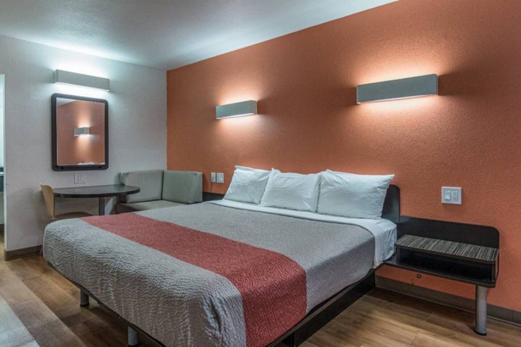 Hotellihuone, jossa on sänky, pieni pöytä, tuoli ja seinään kiinnitetty valaisin.