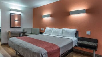 Hotellihuone, jossa on sänky, pieni pöytä, tuoli ja seinään kiinnitetty valaisin.