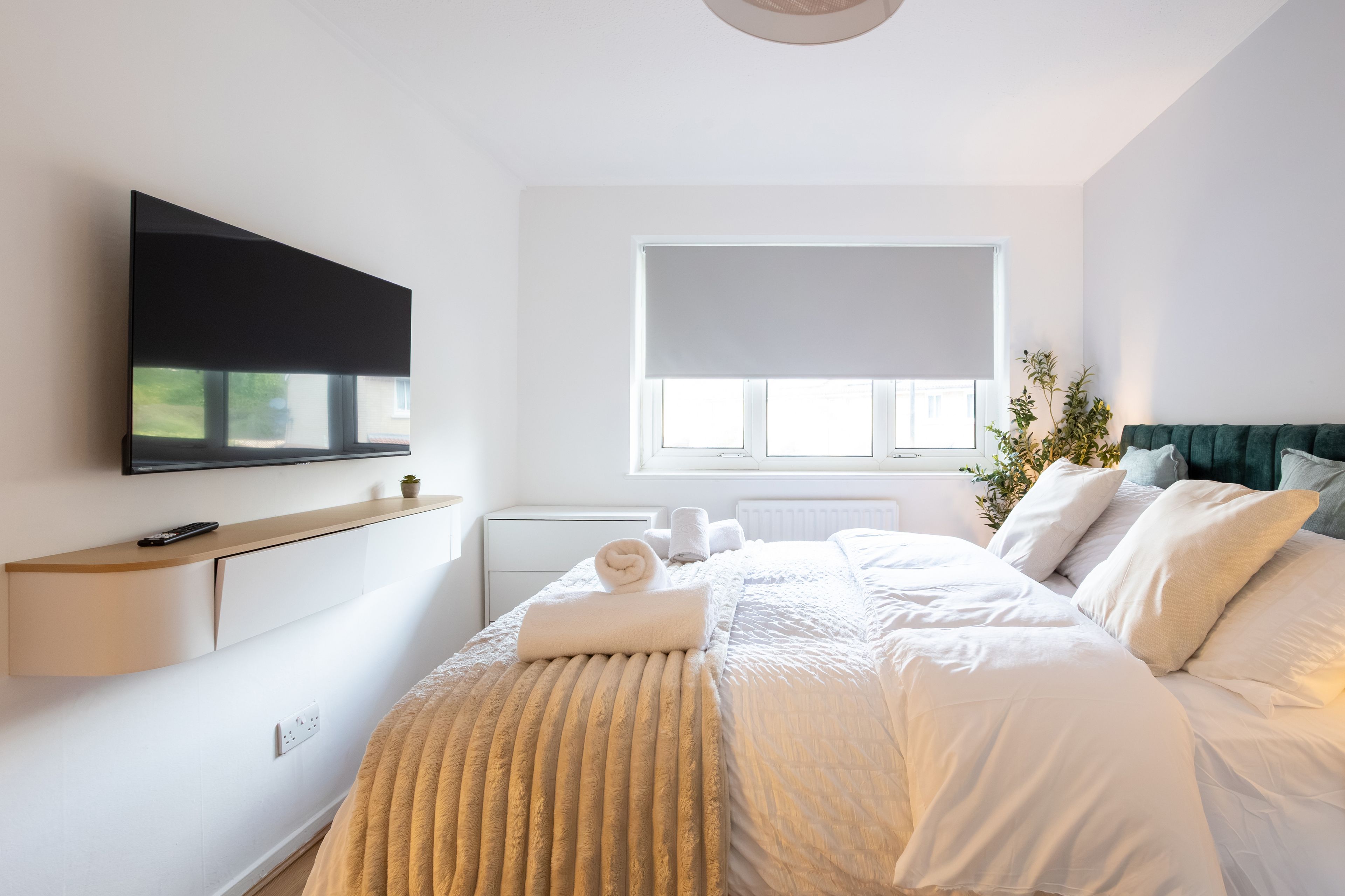 A modern bedroom with a flat-screen TV mounted on the wall, a bed with white bedding, a bedside table with a remote control, and a window with a white blind.
