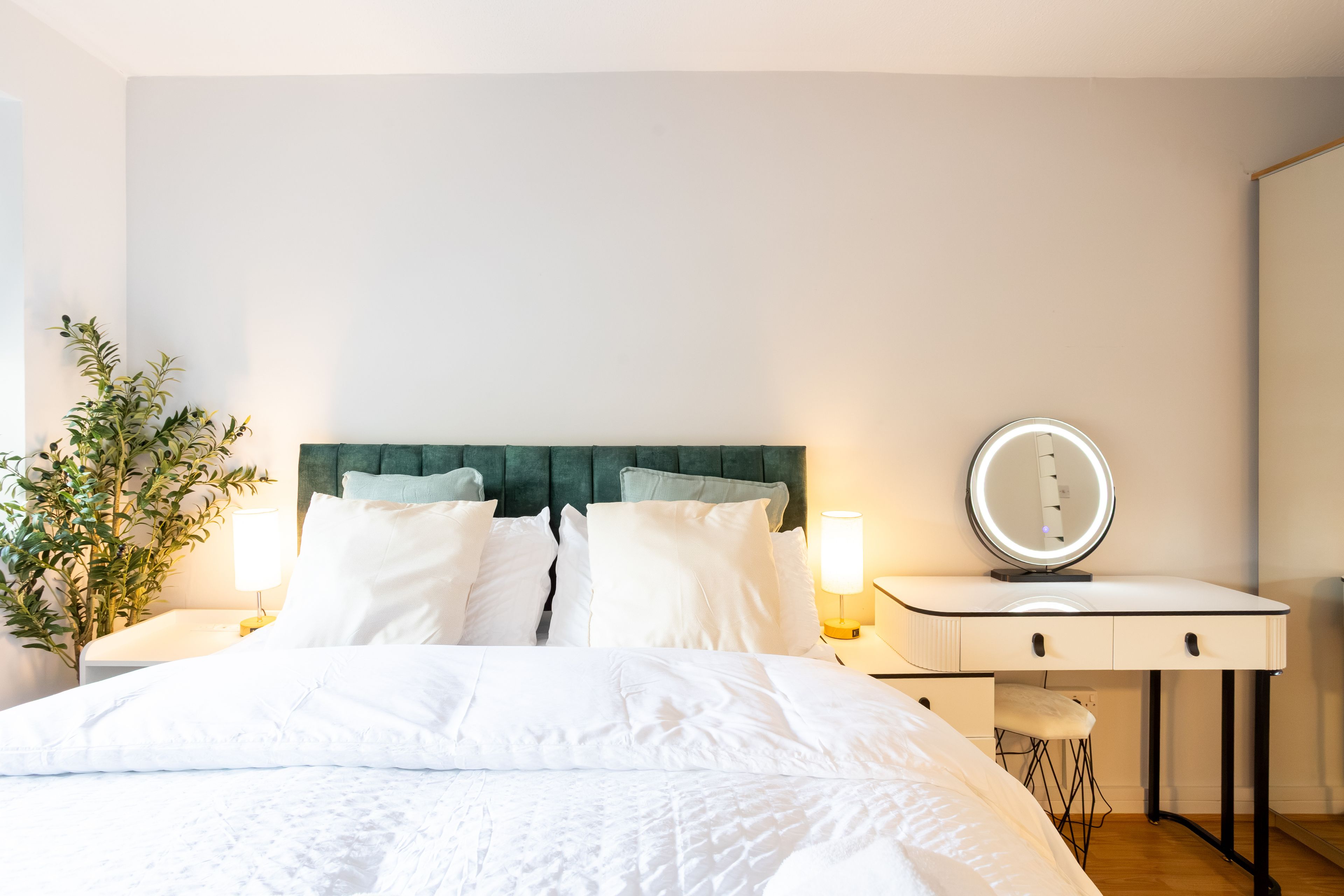 Un lit bien fait, avec du linge de lit blanc, une tête de lit recouverte d’un tissu vert et une table de chevet surmontée d’un miroir rond.